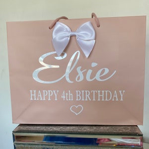 May include: A blush pink gift bag with a white satin bow and matching rope handles. The bag features the name "Elsie" in silver script, along with "HAPPY 4th BIRTHDAY" and a heart symbol.