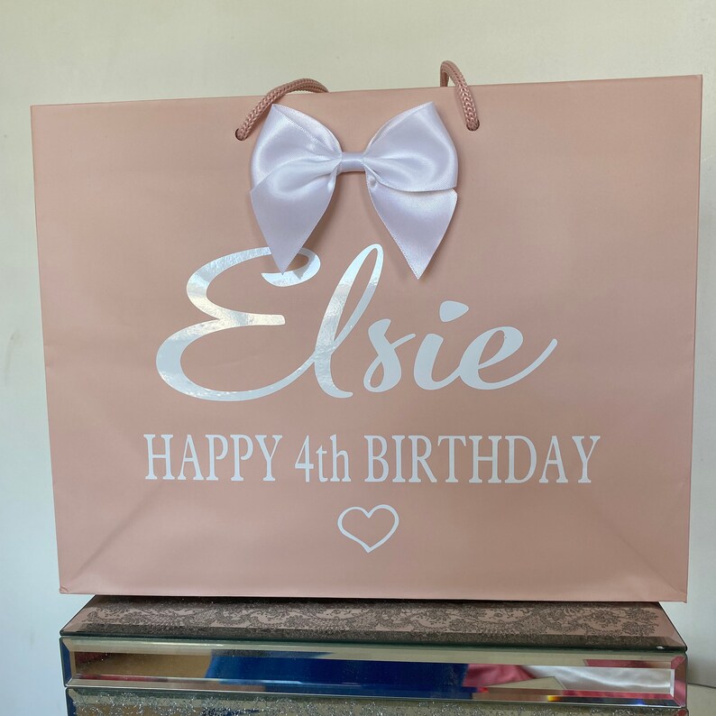 May include: A blush pink gift bag with a white satin bow and matching rope handles. The bag features the name "Elsie" in silver script, along with "HAPPY 4th BIRTHDAY" and a heart symbol.
