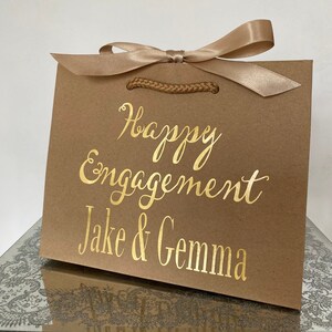 May include: A brown gift bag with a gold satin ribbon and the words "Happy Engagement Jake & Gemma" in gold script. The bag has a braided handle and is sitting on a reflective surface.
