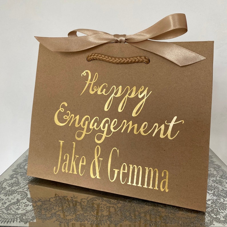 May include: A brown gift bag with a gold satin ribbon and the words "Happy Engagement Jake & Gemma" in gold script. The bag has a braided handle and is sitting on a reflective surface.