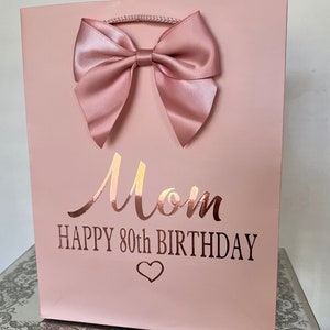 May include: A pink gift bag with a large satin bow. The bag has the word "Mom" in a metallic script, with "HAPPY 80TH BIRTHDAY" below, and a heart symbol. The bag has a rope handle.