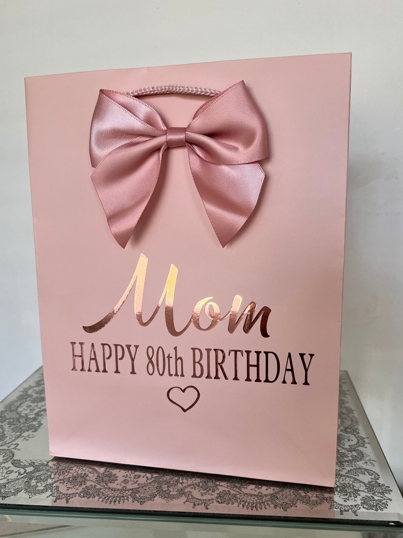 May include: A pink gift bag with a large satin bow. The bag has the word "Mom" in a metallic script, with "HAPPY 80TH BIRTHDAY" below, and a heart symbol. The bag has a rope handle.