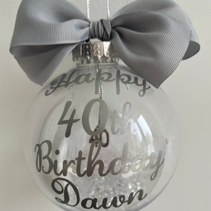 May include: A clear glass bauble with silver text reading "Happy 40th Birthday Dawn." The ornament is decorated with a grey bow and filled with white decorative material. A silver string is attached for hanging.