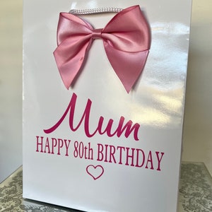 May include: A white gift bag with a large pink satin bow and a white cord handle. The bag has the words "Mum HAPPY 80th BIRTHDAY" printed in pink, with a heart symbol below the text. The bag is glossy.