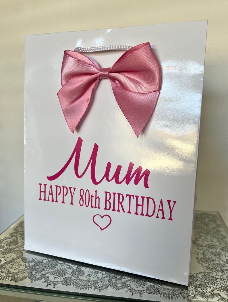 May include: A white gift bag with a large pink satin bow and a white cord handle. The bag has the words "Mum HAPPY 80th BIRTHDAY" printed in pink, with a heart symbol below the text. The bag is glossy.