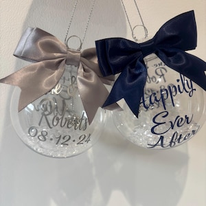 May include: Two clear baubles with silver bows and hanging loops. One has a silver bow and the text "Di Roberts 08.12.24". The other has a navy blue bow and the text "Happily Ever After". Both are filled with small, clear beads.