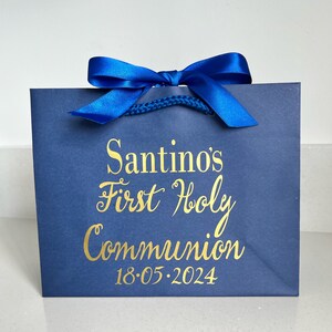 Pode incluir: Uma sacola de presente azul marinho com um la&ccedil;o de fita de cetim azul royal. A escrita dourada diz "Santino's First Holy Communion 18.05.2024". A sacola tem uma al&ccedil;a de cord&atilde;o azul torcido.
