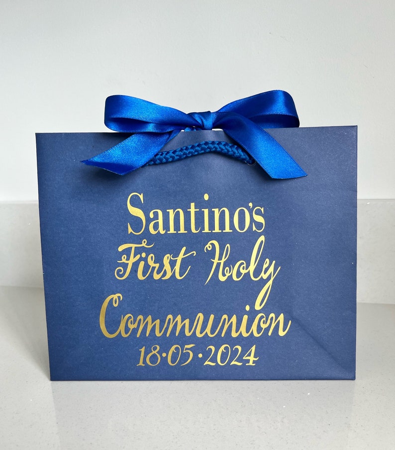 Pode incluir: Uma sacola de presente azul marinho com um la&ccedil;o de fita de cetim azul royal. A escrita dourada diz "Santino's First Holy Communion 18.05.2024". A sacola tem uma al&ccedil;a de cord&atilde;o azul torcido.