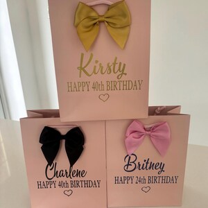 May include: Pink gift bags with decorative bows in gold, black, and pink. Each bag has a name and birthday message printed on it. The bags are for a 40th and 24th birthday.