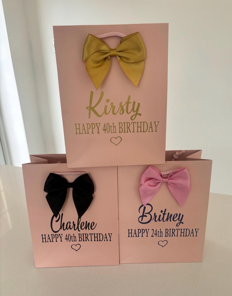 May include: Pink gift bags with decorative bows in gold, black, and pink. Each bag has a name and birthday message printed on it. The bags are for a 40th and 24th birthday.