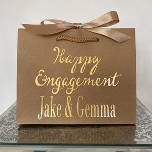 May include: A brown paper gift bag with a gold satin ribbon and rope handles. The words "Happy Engagement" and the names "Jake & Gemma" are printed in gold script on the front of the bag.