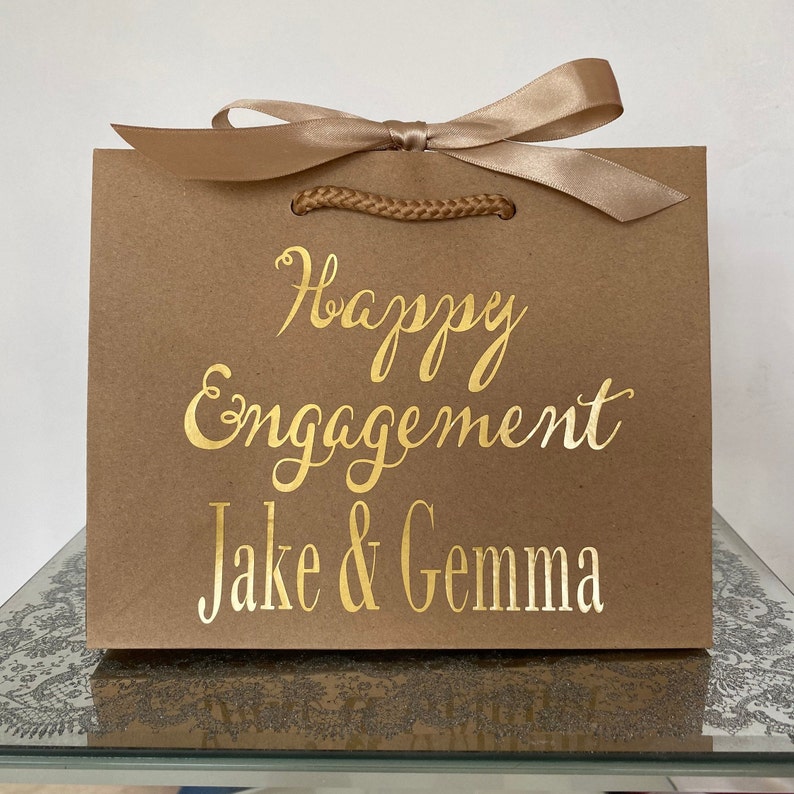 May include: A brown paper gift bag with a gold satin ribbon and rope handles. The words "Happy Engagement" and the names "Jake & Gemma" are printed in gold script on the front of the bag.