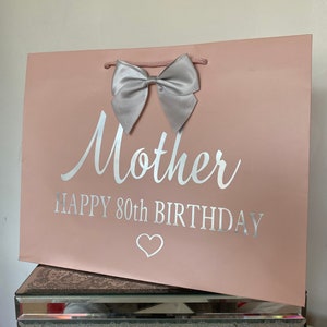 May include: A pink gift bag with a silver bow and the words "Mother HAPPY 80th BIRTHDAY" in silver lettering. A white heart is below the text. The bag has a pink cord handle.