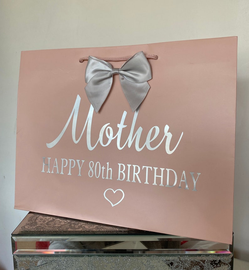 May include: A pink gift bag with a silver bow and the words "Mother HAPPY 80th BIRTHDAY" in silver lettering. A white heart is below the text. The bag has a pink cord handle.