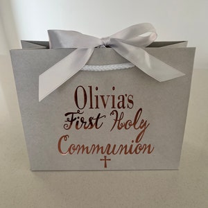Pode incluir: Uma sacola de presente cinza amarrada com um la&ccedil;o de fita prateada. A sacola tem o texto "Olivia's First Holy Communion" em escrita cor de cobre, com uma pequena cruz abaixo. A sacola &eacute; projetada para uma celebra&ccedil;&atilde;o religiosa.