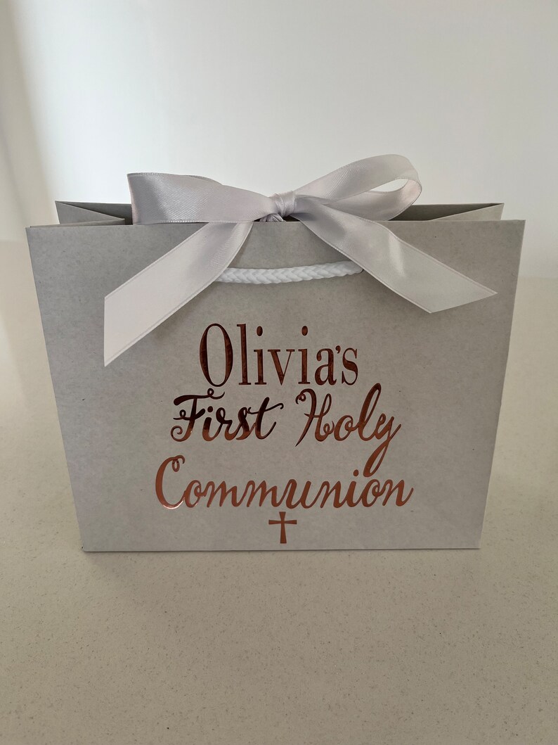 Pode incluir: Uma sacola de presente cinza amarrada com um la&ccedil;o de fita prateada. A sacola tem o texto "Olivia's First Holy Communion" em escrita cor de cobre, com uma pequena cruz abaixo. A sacola &eacute; projetada para uma celebra&ccedil;&atilde;o religiosa.
