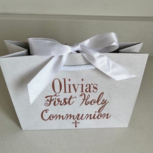 Pode incluir: Uma sacola de presente cinza claro amarrada com uma fita de cetim branca. A sacola tem o texto "Olivia's First Holy Communion" em script marrom. Uma pequena cruz est&aacute; abaixo do texto. A sacola tem uma al&ccedil;a de corda branca.