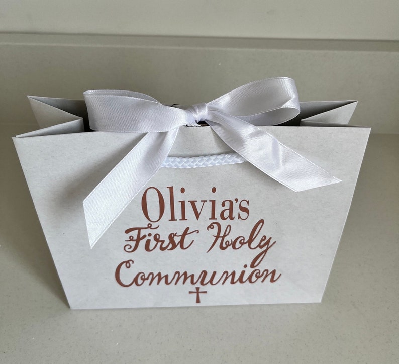Pode incluir: Uma sacola de presente cinza claro amarrada com uma fita de cetim branca. A sacola tem o texto "Olivia's First Holy Communion" em script marrom. Uma pequena cruz est&aacute; abaixo do texto. A sacola tem uma al&ccedil;a de corda branca.