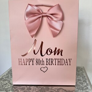 May include: A light pink gift bag with a large satin bow. The bag has the words "Mom HAPPY 80th BIRTHDAY" in a metallic rose gold font, with a heart symbol below the text. A twisted cord handle is at the top.