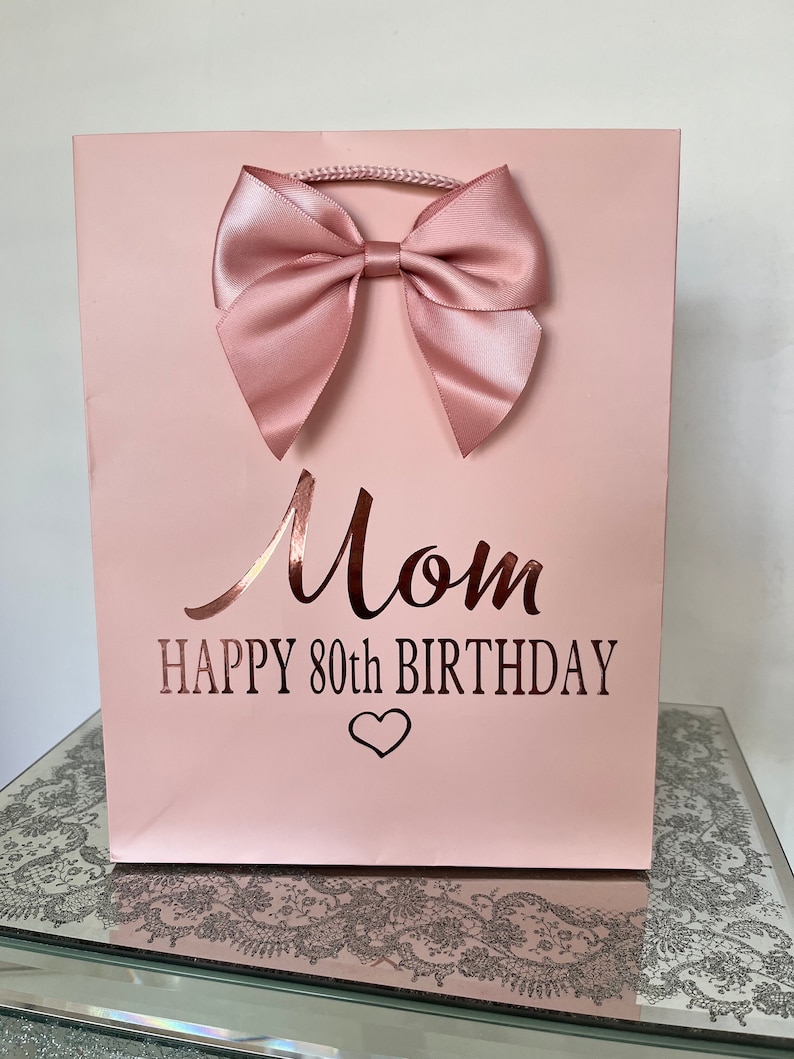 May include: A light pink gift bag with a large satin bow. The bag has the words "Mom HAPPY 80th BIRTHDAY" in a metallic rose gold font, with a heart symbol below the text. A twisted cord handle is at the top.