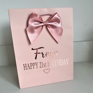 May include: A pink gift bag with a large satin bow at the top. The bag has the name "Freya" and the text "HAPPY 21st BIRTHDAY" in rose gold lettering, with a heart symbol below.