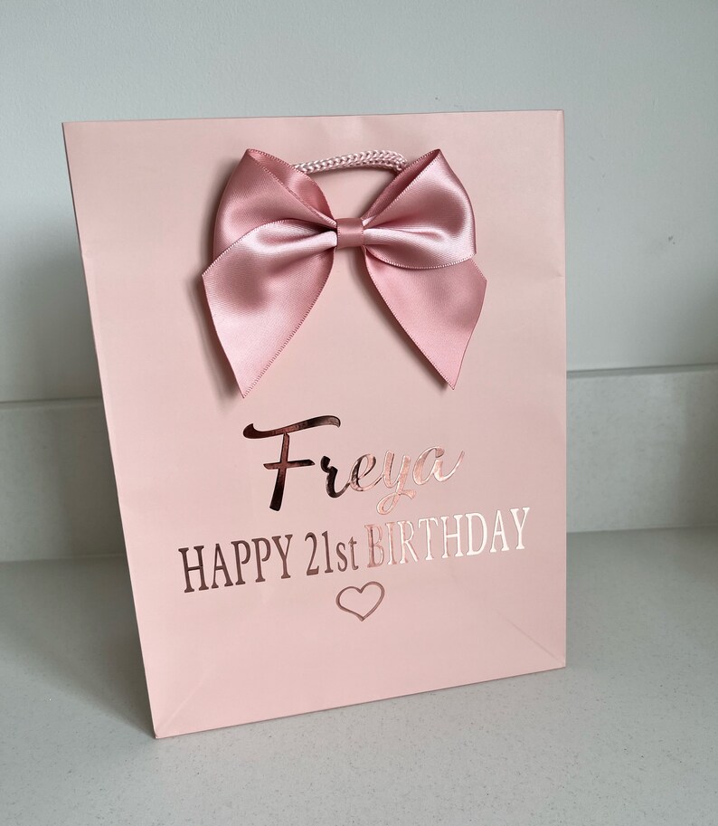 May include: A pink gift bag with a large satin bow at the top. The bag has the name "Freya" and the text "HAPPY 21st BIRTHDAY" in rose gold lettering, with a heart symbol below.