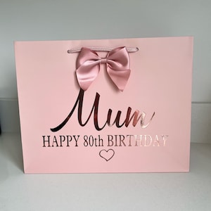 May include: A pale pink gift bag with a satin bow and rope handles. The bag features the word "Mum" in a cursive font, along with "HAPPY 80th BIRTHDAY" and a heart symbol, in a metallic rose gold colour.
