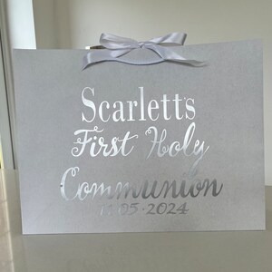 Pode incluir: Uma sacola de presente cinza claro com uma fita de cetim branca na parte superior. A sacola apresenta texto prateado que diz "Scarlett's First Holy Communion" e a data "11.05.2024".