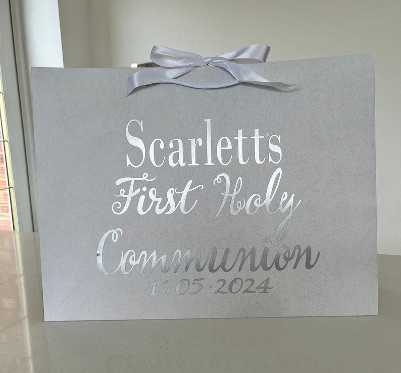 Pode incluir: Uma sacola de presente cinza claro com uma fita de cetim branca na parte superior. A sacola apresenta texto prateado que diz "Scarlett's First Holy Communion" e a data "11.05.2024".