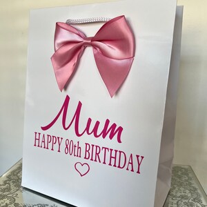 May include: White gift bag with a pink satin bow and a white cord handle. The bag has the words "Mum HAPPY 80th BIRTHDAY" in pink script. A pink heart is below the text.