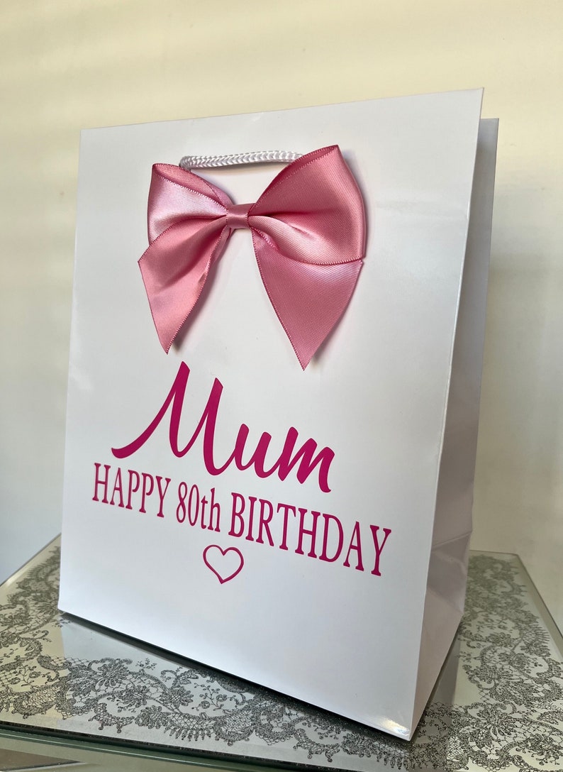 May include: White gift bag with a pink satin bow and a white cord handle. The bag has the words "Mum HAPPY 80th BIRTHDAY" in pink script. A pink heart is below the text.