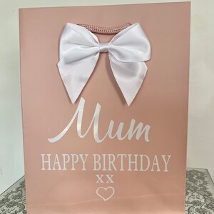 May include: A pink gift bag with a white satin bow and a woven handle. The bag has the words "Mum HAPPY BIRTHDAY" printed in white, along with two "XX" symbols and a heart.