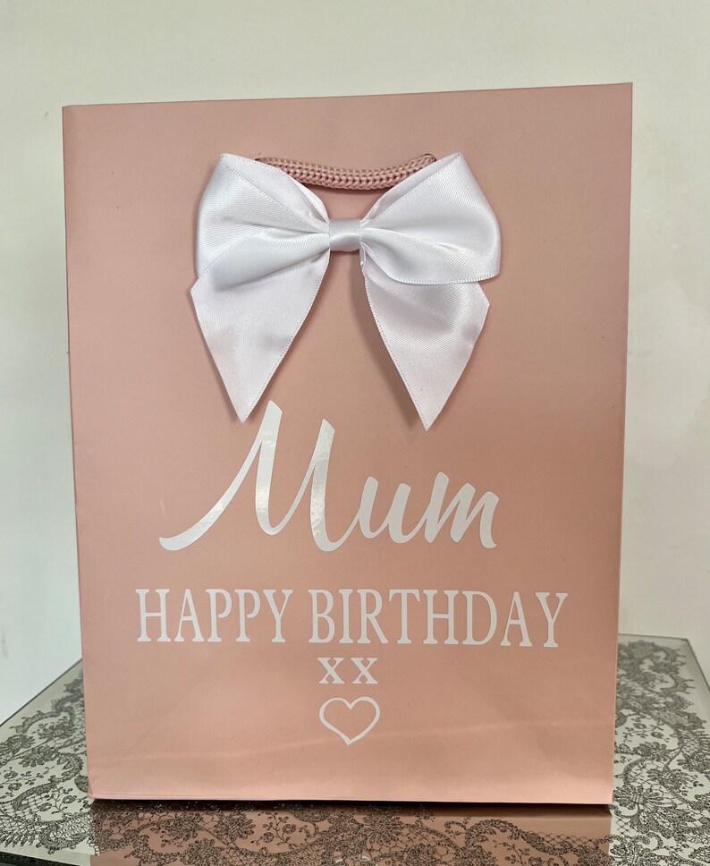 May include: A pink gift bag with a white satin bow and a woven handle. The bag has the words "Mum HAPPY BIRTHDAY" printed in white, along with two "XX" symbols and a heart.