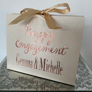 May include: A cream-colored gift bag with a gold satin ribbon tied in a bow. The words "Happy Engagement Gemma & Michelle" are printed in a rose gold script. The bag has white rope handles.