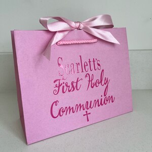 Pode incluir: Uma sacola de presente rosa com la&ccedil;o de fita de cetim e al&ccedil;as de corda. A sacola apresenta o texto "Scarlett's First Holy Communion" em rosa met&aacute;lico, junto com um s&iacute;mbolo de cruz.