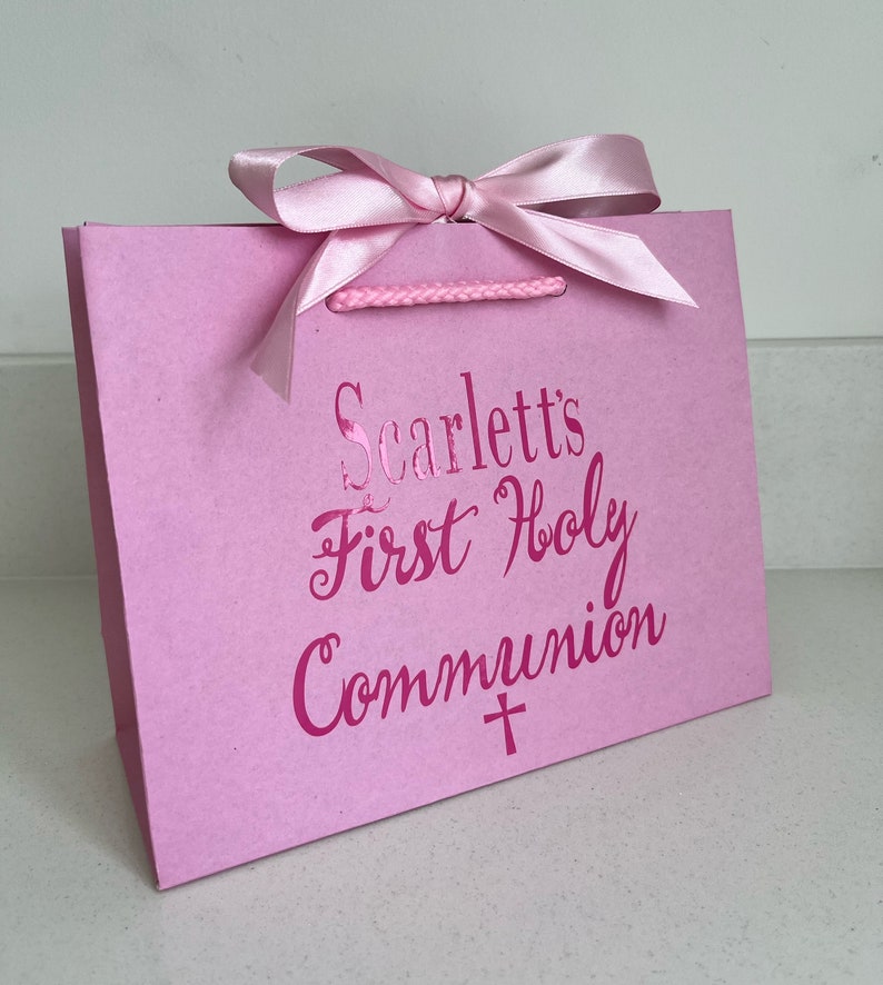 Pode incluir: Uma sacola de presente rosa com la&ccedil;o de fita de cetim e al&ccedil;as de corda. A sacola apresenta o texto "Scarlett's First Holy Communion" em rosa met&aacute;lico, junto com um s&iacute;mbolo de cruz.