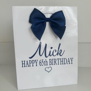 May include: White gift bag with a navy blue satin bow and a white cord handle. The bag has the name "Mick", the text "HAPPY 65TH BIRTHDAY", and a heart symbol printed in navy blue.