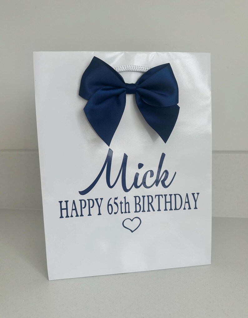 May include: White gift bag with a navy blue satin bow and a white cord handle. The bag has the name "Mick", the text "HAPPY 65TH BIRTHDAY", and a heart symbol printed in navy blue.