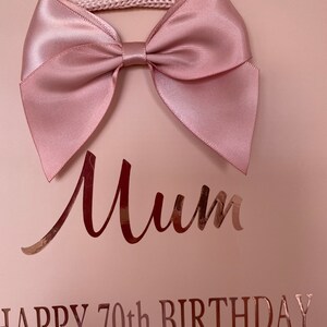 May include: A light pink gift bag with a large satin bow at the top. The bag has the word "Mum" in a cursive font and the text "HAPPY 70th BIRTHDAY" in a metallic rose gold color.
