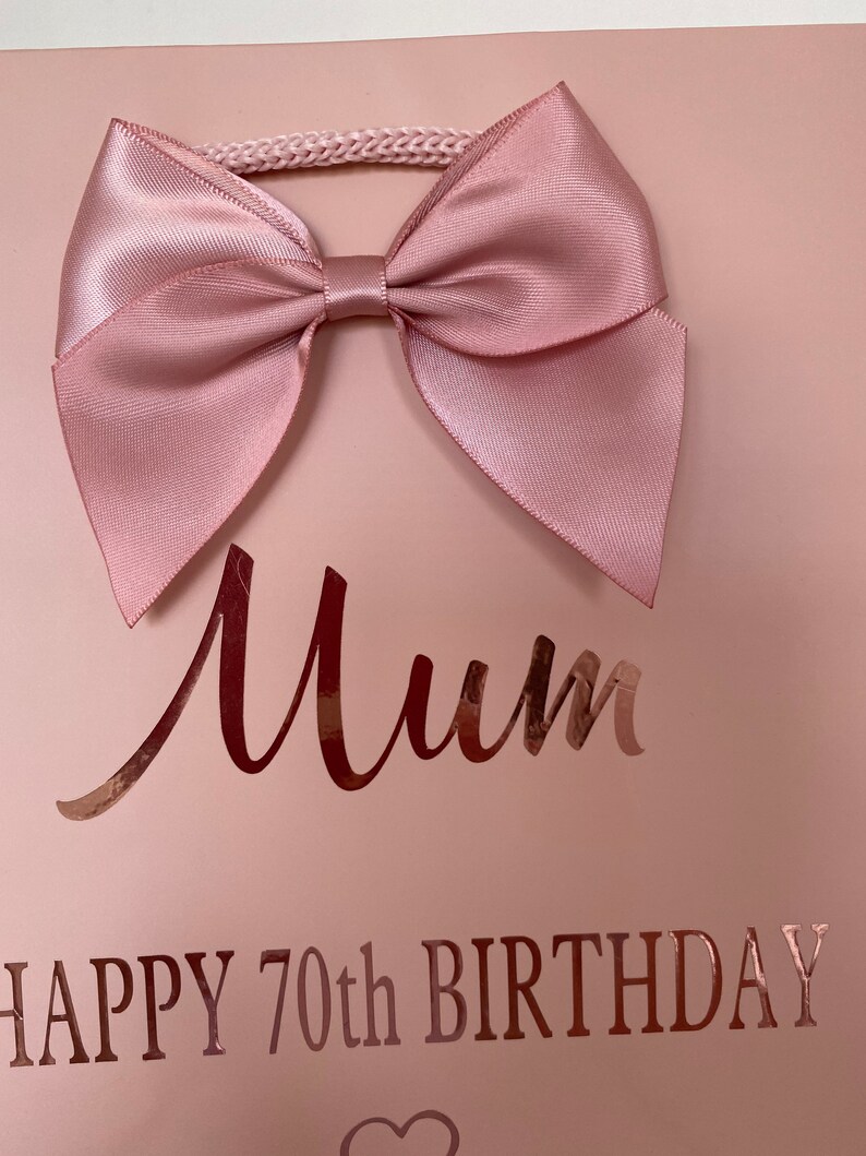 May include: A light pink gift bag with a large satin bow at the top. The bag has the word "Mum" in a cursive font and the text "HAPPY 70th BIRTHDAY" in a metallic rose gold color.