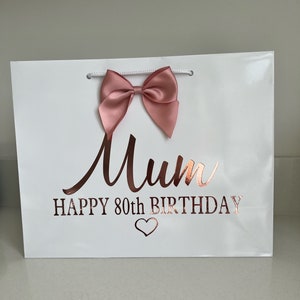 May include: White gift bag with a pink satin bow and rose gold text that reads "Mum HAPPY 80th BIRTHDAY" with a heart symbol. The bag has white rope handles.