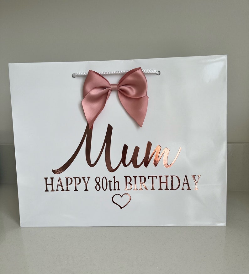 May include: White gift bag with a pink satin bow and rose gold text that reads "Mum HAPPY 80th BIRTHDAY" with a heart symbol. The bag has white rope handles.