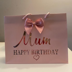 May include: A pink gift bag with a large bow and the words "Mum HAPPY BIRTHDAY" in a metallic rose gold font. The bag has matching pink rope handles and a heart symbol.
