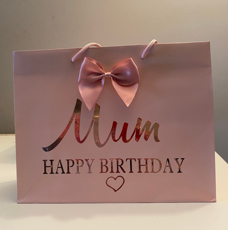 May include: A pink gift bag with a large bow and the words "Mum HAPPY BIRTHDAY" in a metallic rose gold font. The bag has matching pink rope handles and a heart symbol.