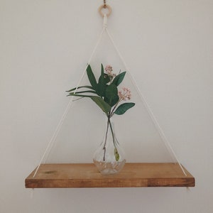 Puede incluir: Estante flotante de madera con un jarrón de vidrio transparente que contiene flores rosas y hojas verdes. El estante está suspendido por una cuerda blanca de un anillo de madera fijado a la pared. El estante es de madera marrón claro.