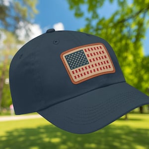 Könnte beinhalten: Marineblaue Baseballkappe mit einem rechteckigen Aufnäher, der eine amerikanische Flagge zeigt. Die Flagge ersetzt die Streifen durch rote Reißzwecken und hat ein blaues Feld mit weißen Sternen. Der Aufnäher ist mit einem braunen Rand eingefasst.