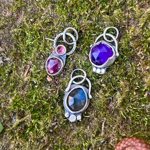 May include: Three silver pendants featuring faceted gemstones. One pendant has a small pink stone and a larger deep red stone. Another features a large, round, faceted purple stone. The third displays a dark, iridescent labradorite stone.