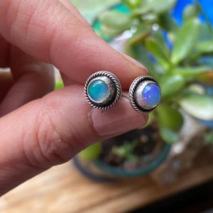 Könnte beinhalten: Silberne Ohrstecker mit Opalsteinen. Jeder Ohrring hat einen runden Opal, der in einer gedrehten, seilartigen Umrandung gefasst ist. Die Opale zeigen eine Mischung aus blauen, grünen und weißen Farben und erzeugen einen schimmernden Effekt.