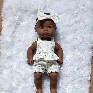 May include: A Black doll wearing a white outfit with a rainbow pattern. The doll is wearing a headband with a rainbow bow. The doll is lying on a white fluffy blanket.