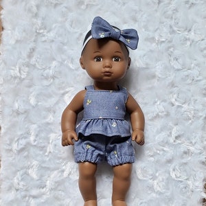 May include: A dark-skinned baby doll wearing a blue denim outfit with white floral accents. The doll has a headband with a blue bow and is lying on a white fluffy surface.
